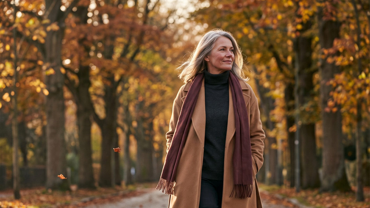 Woman in a camel wool coat and burgundy scarf walking a tree-lined avenue in autumn